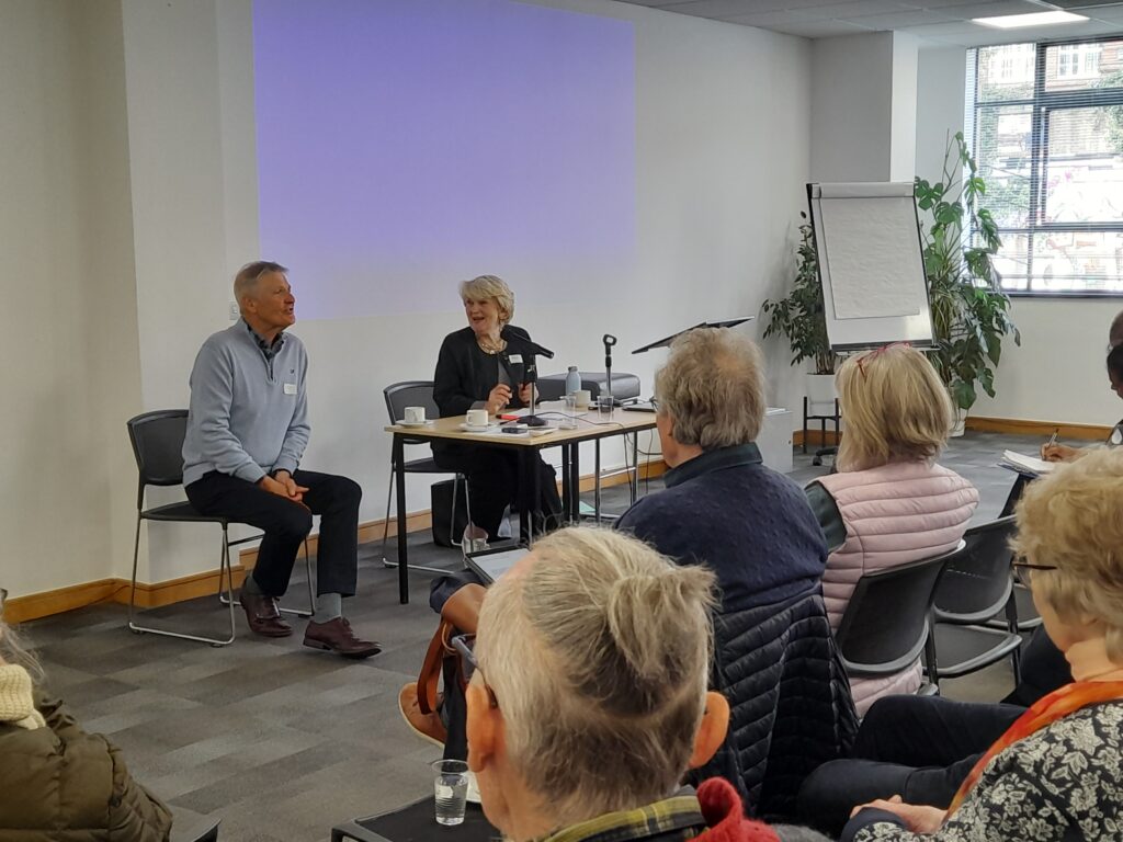 David from Our Life and Our Choice and Mary from Camphill Families and Friends are sitting behind a table at the front of the meeting room. David is answering a question from the audience. The backs of the audience's heads are visible.